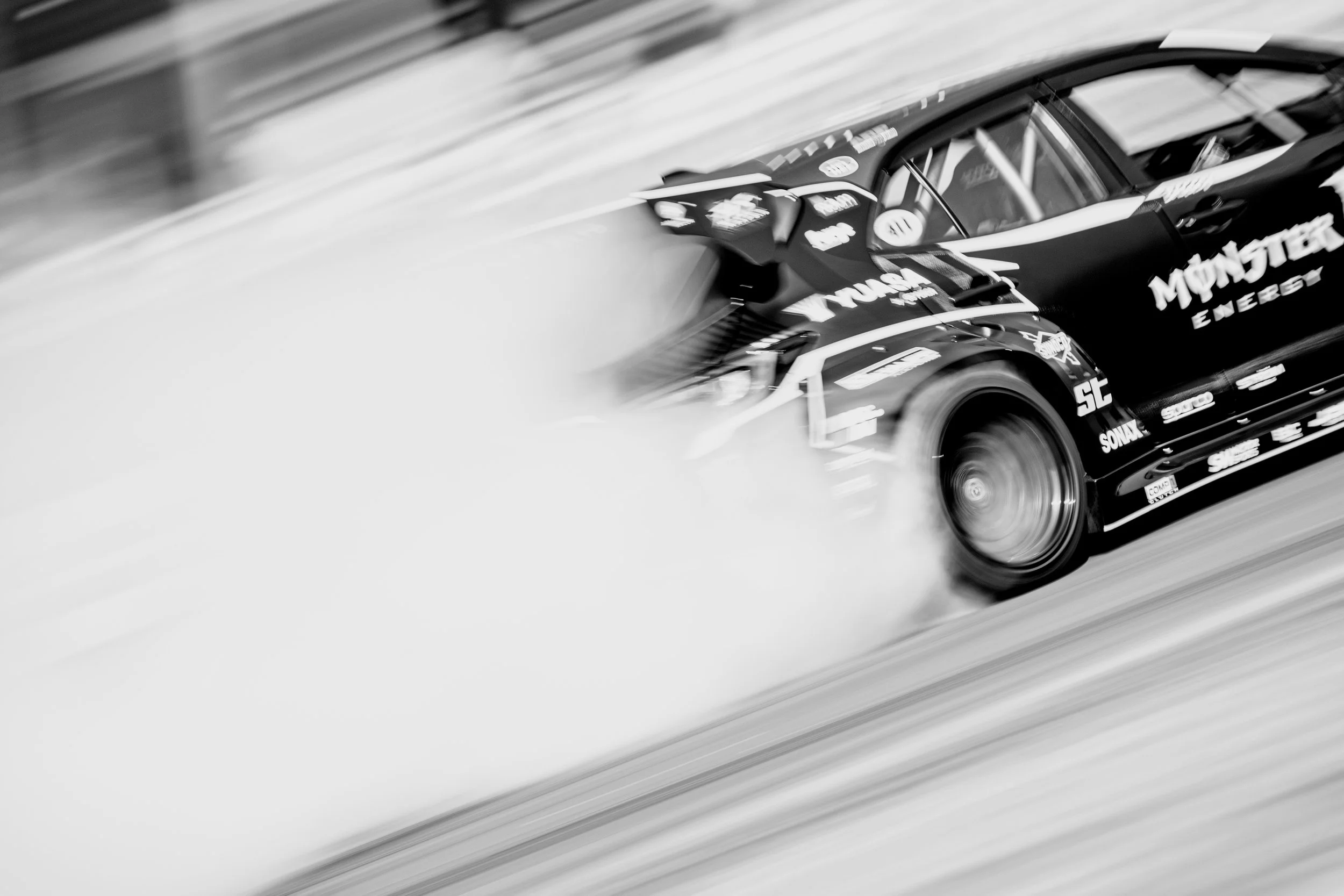 Black race car with sponsor logos racing on a track, kicking up dust, moving at high speed, in black and white.