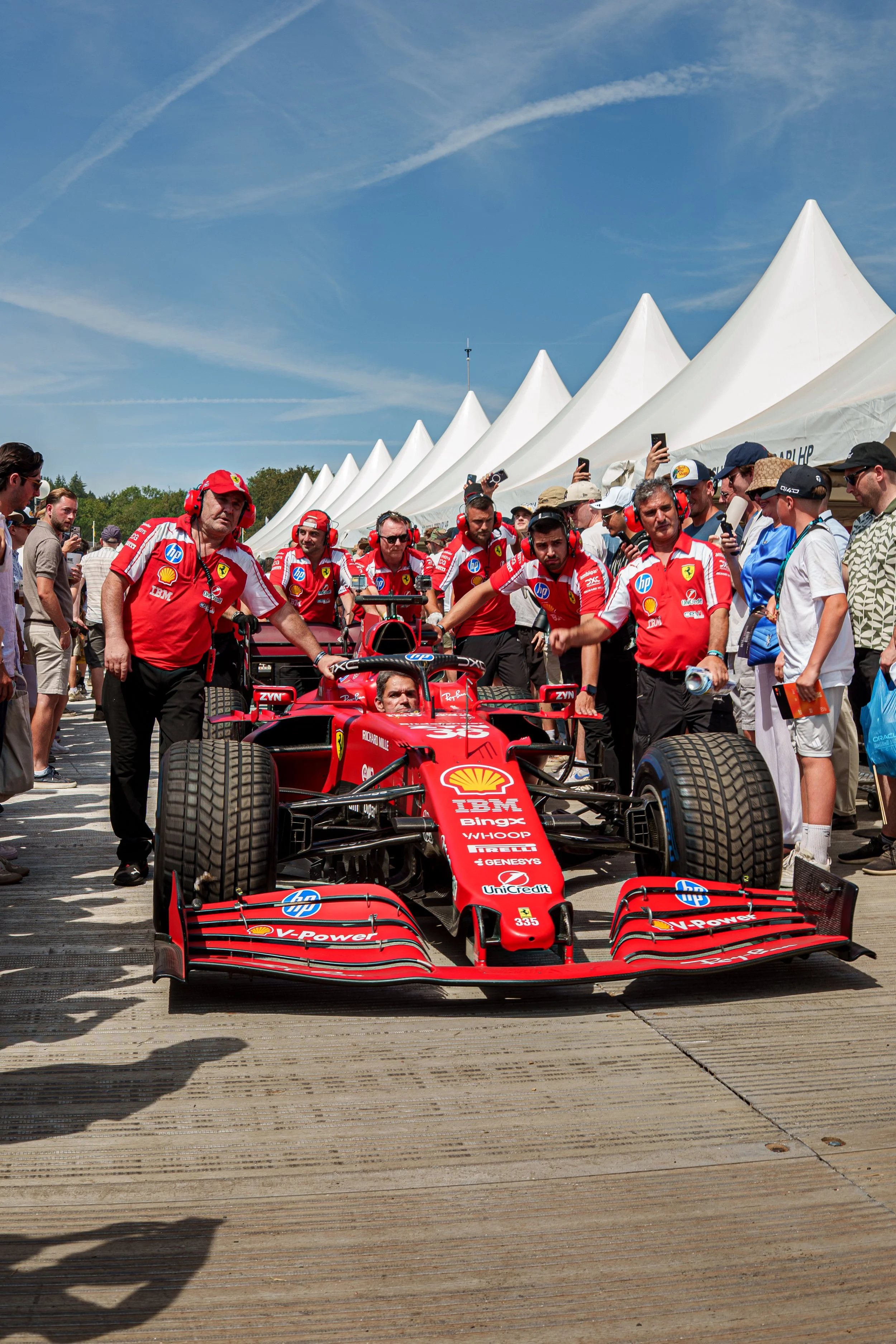 A red Formula 1 race car being pushed by a group of team members in red uniforms and ear protection, surrounded by onlookers, under a row of white tents on a sunny day.