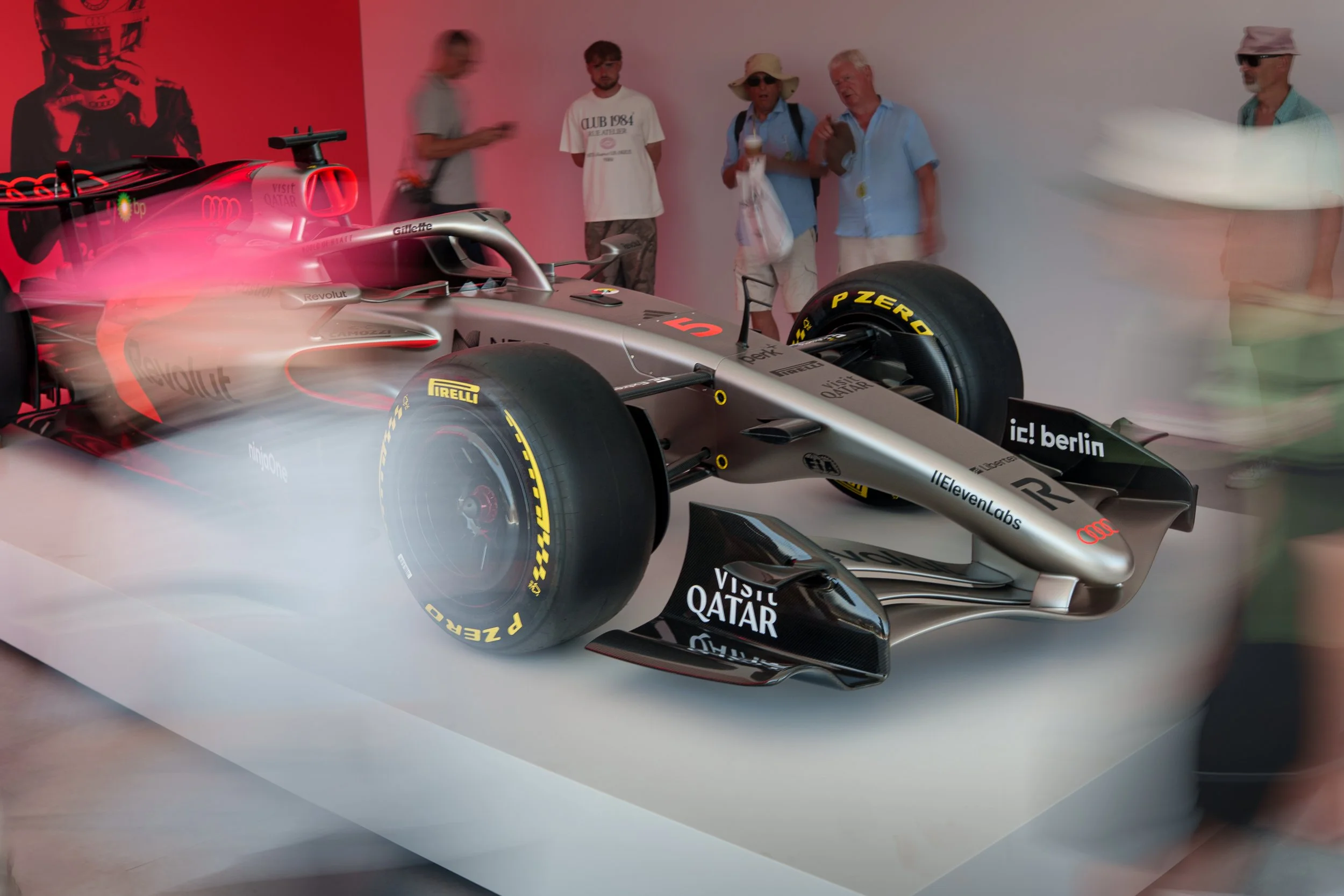 A red and silver Formula 1 race car on display with blurred visitors in the background at an indoor exhibition.