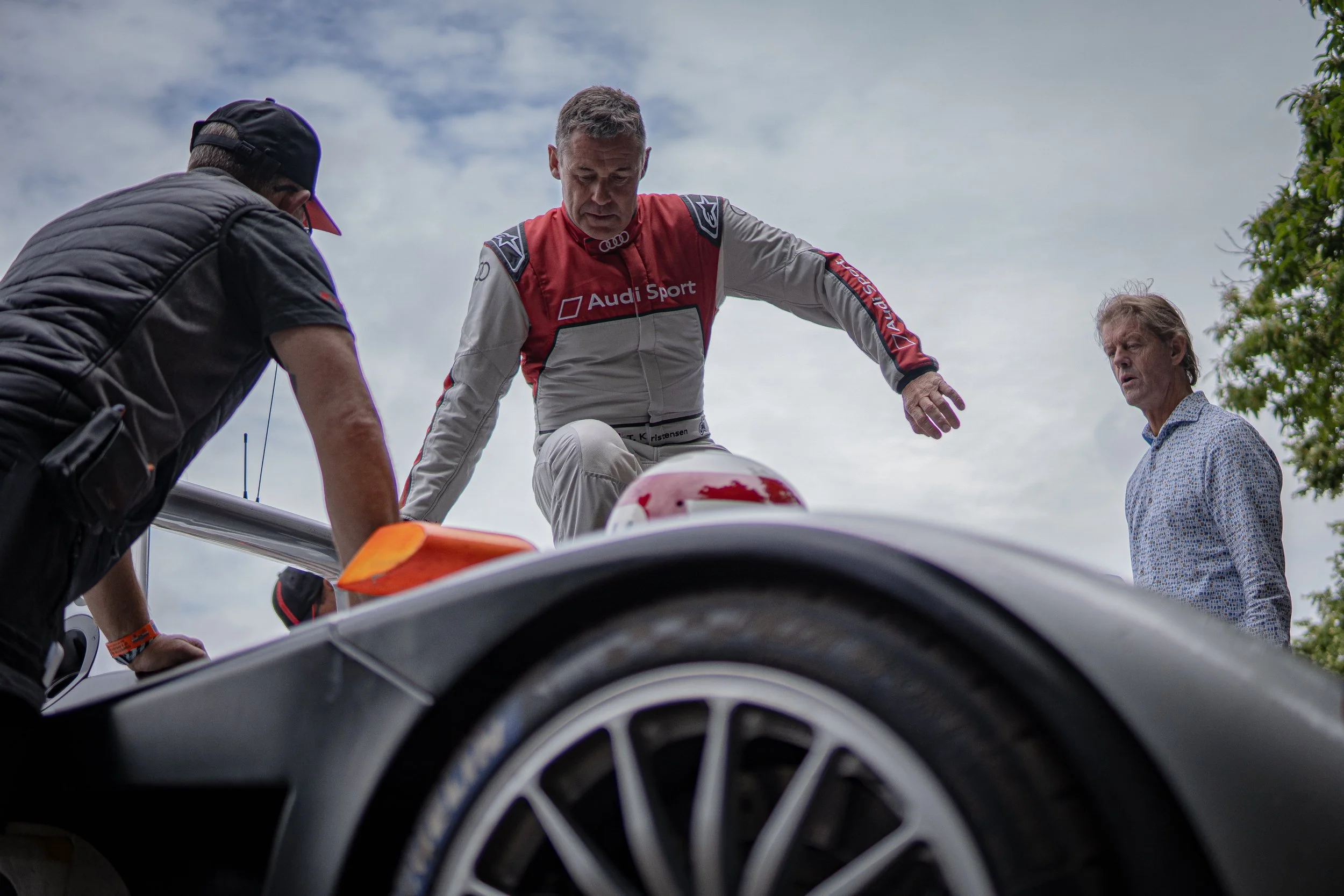 Race car driver in a red suit with sponsorship logos, standing next to a race car, with team members and officials nearby, during a racing event under a cloudy sky.