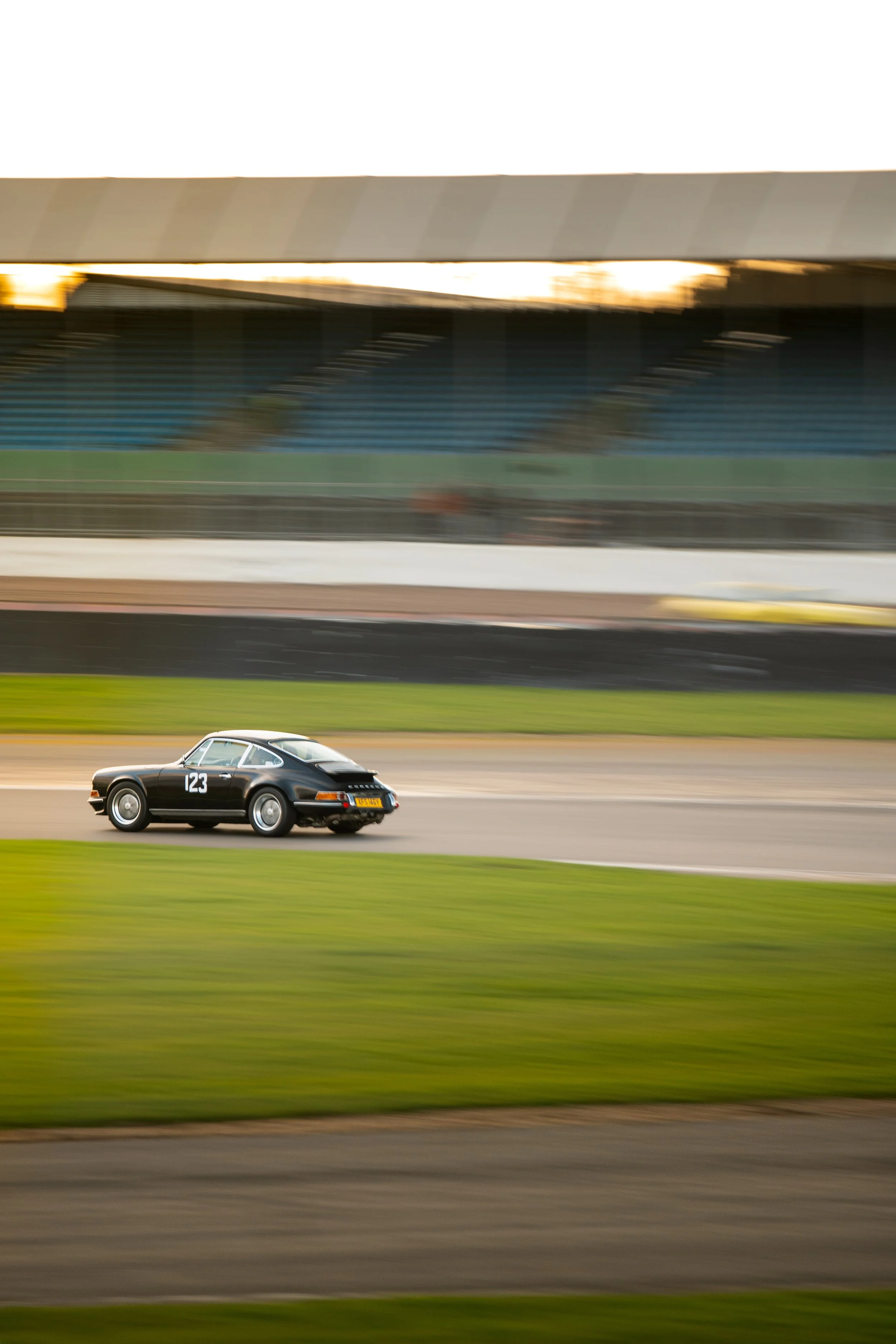 A black vintage car with the number 123 on the side driving at the Pomeroy Trophy at Silverstone during sunset, with a blurred background of green grass and grandstands.