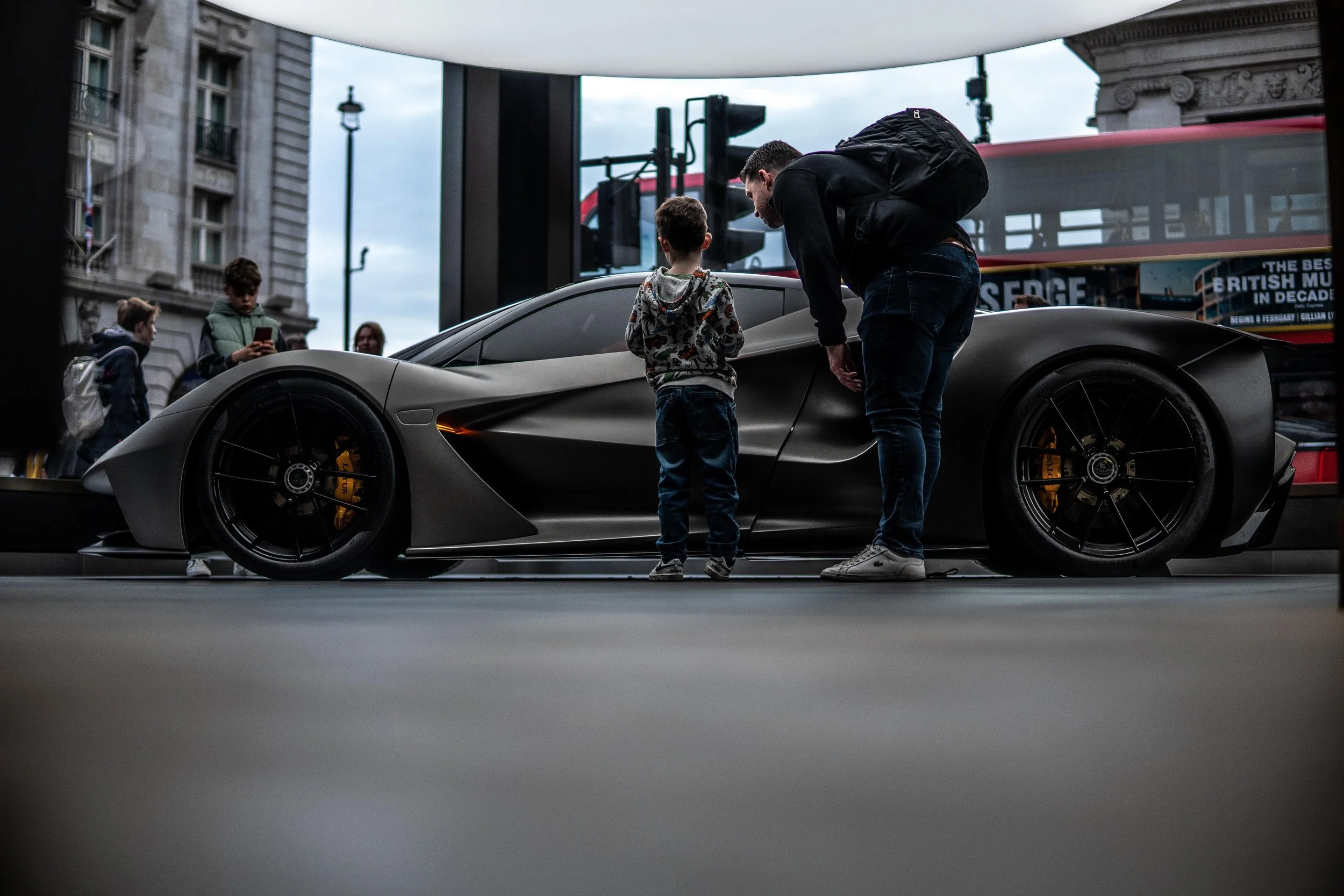A man and a boy are standing in front of a silver sports car, possibly a Lotus, on a city street. The man is leaning toward the boy as they look at the car. In the background, there is a red double-decker bus and several children, one of whom is holding a phone, with city buildings and a streetlamp.