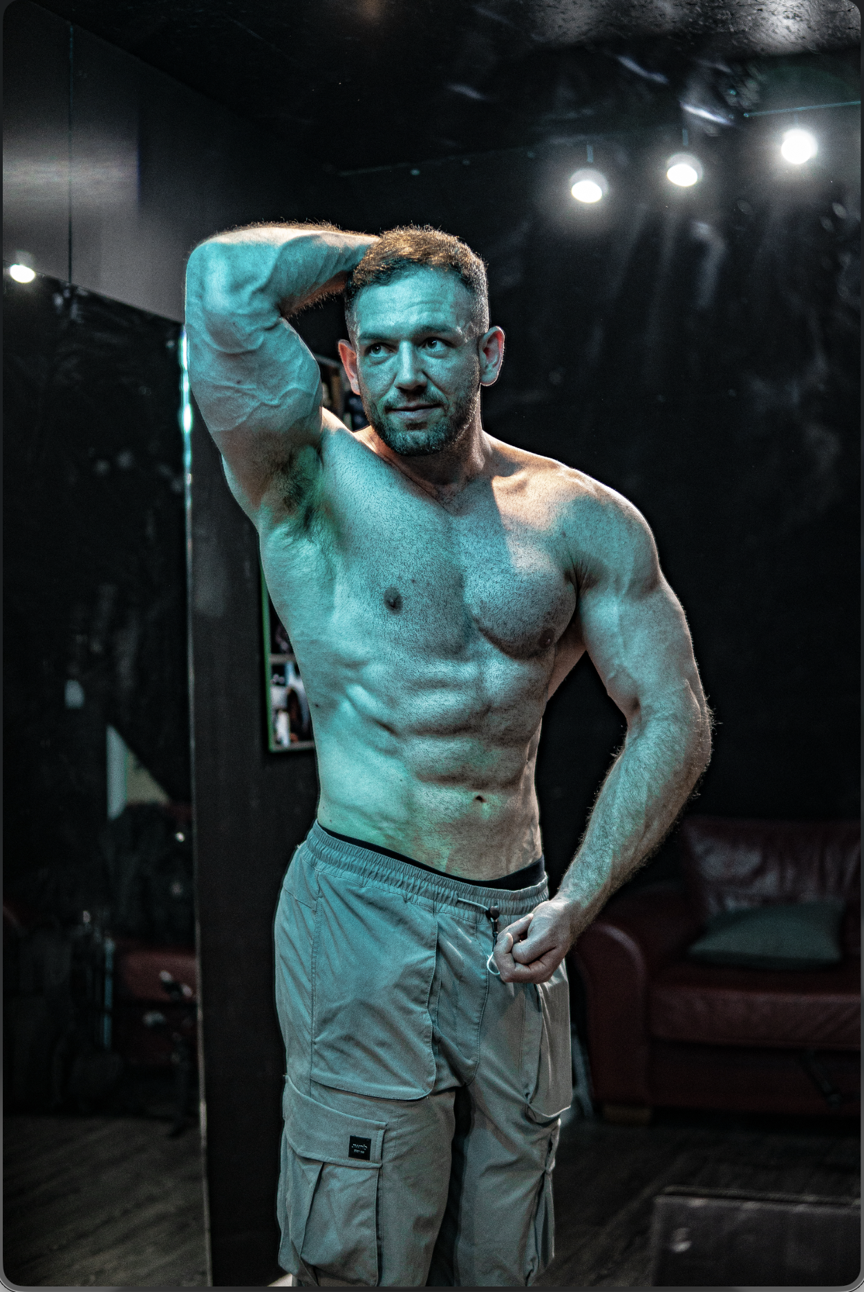 A muscular, bodybuilder with short hair and a beard standing with one arm raised behind his head and the other hand resting on his waist in a dimly lit room with black walls and ceiling, and bright stage lights overhead.