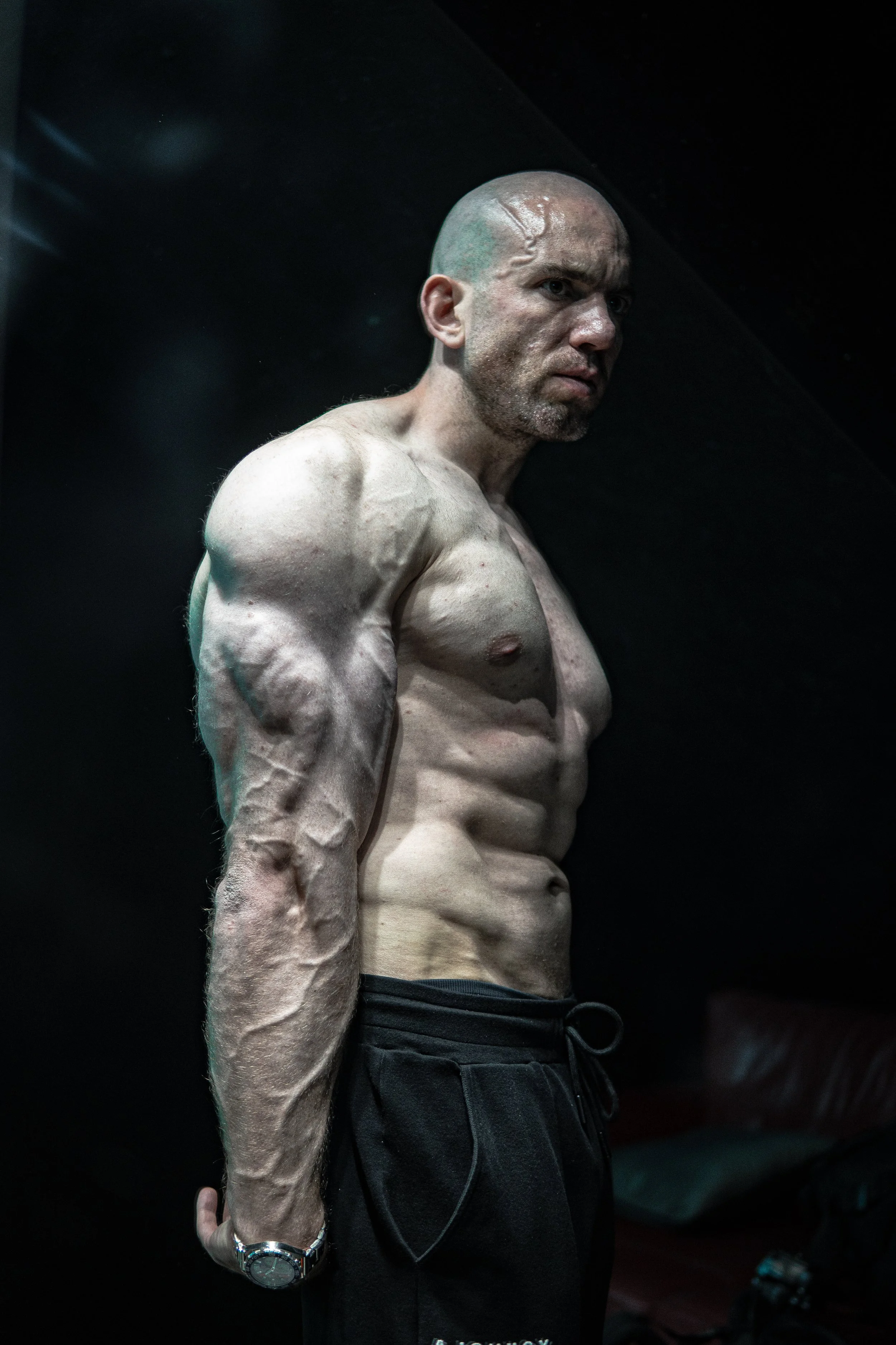 A shirtless bodybuilder with a shaved head and muscular physique, standing against a dark background, wearing black shorts and a silver wristwatch.