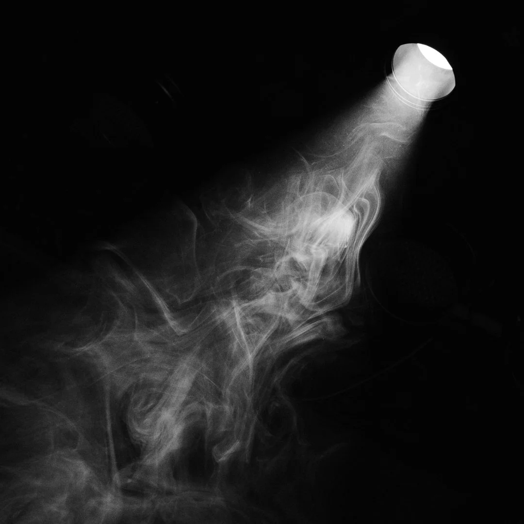 A spotlight cutting through smoke in darkness, representing stepping into visibility and finding your voice.