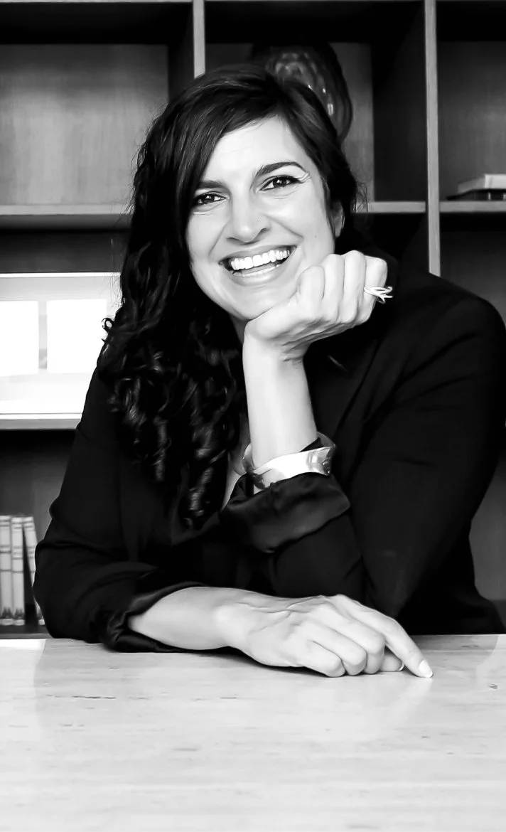 Black and white photo of a woman with dark, wavy hair smiling, resting her chin on her hand, sitting at a desk in front of a bookshelf.