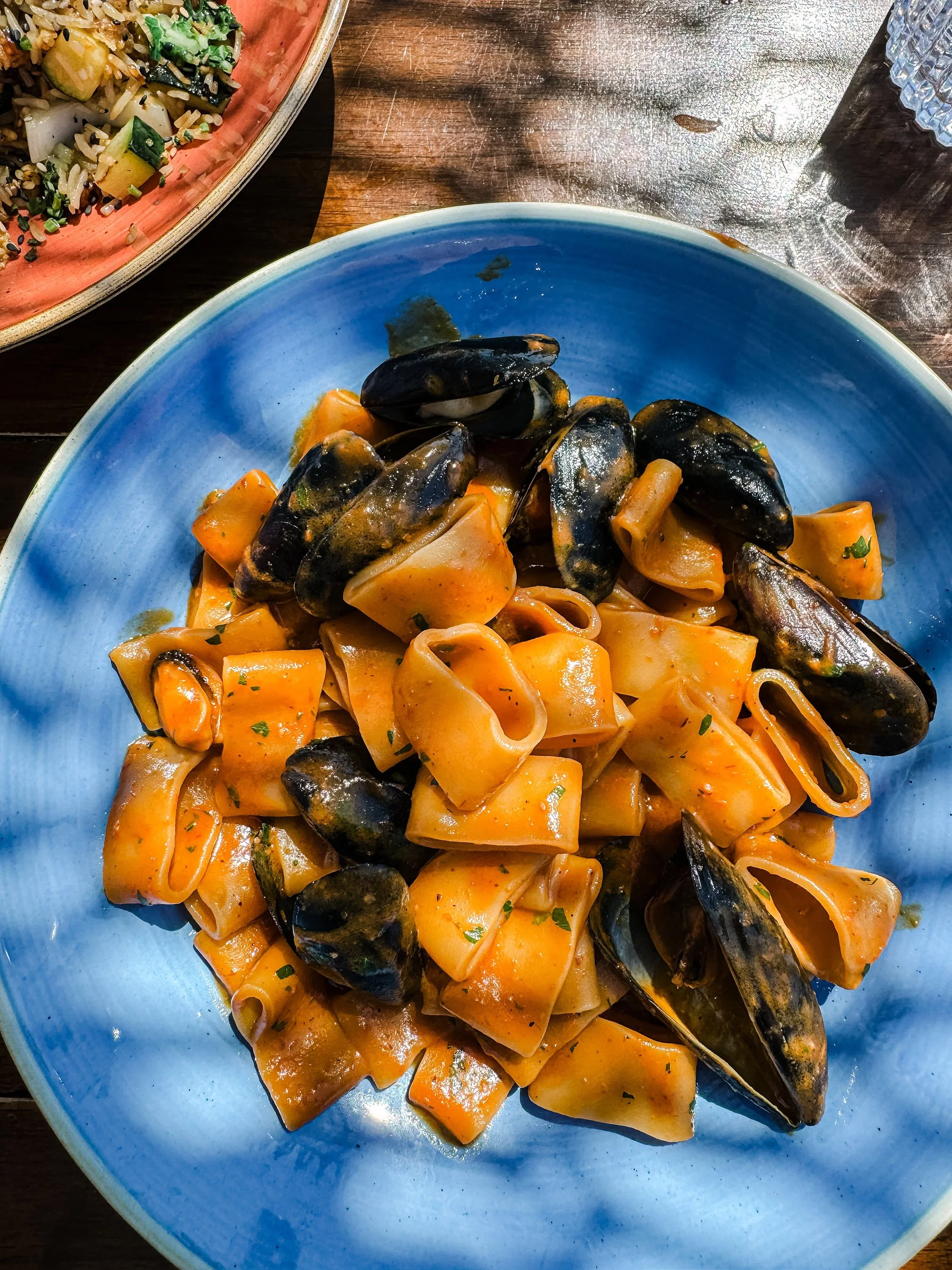 A blue plate of pasta with mussels, chopped tomatoes, and herbs on a wooden table.