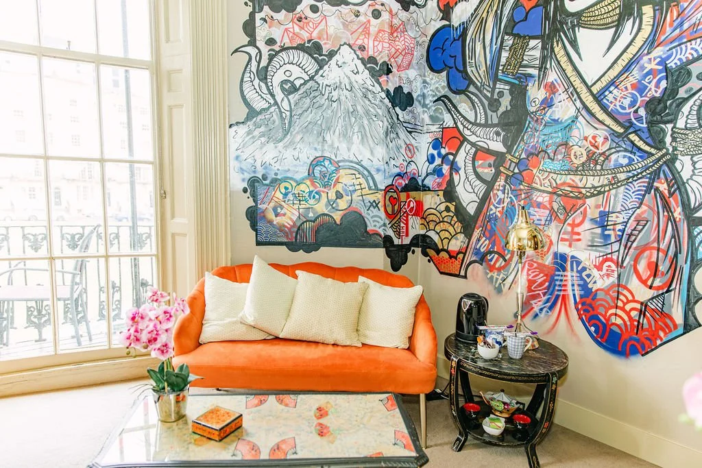 A cozy living room corner with an orange sofa, three beige pillows, a marble-top side table with a kettle, cups, and snacks, a small potted pink flower, and a large colorful mural with Mount Fuji, clouds, and abstract patterns on the wall next to a glass door leading to a balcony.