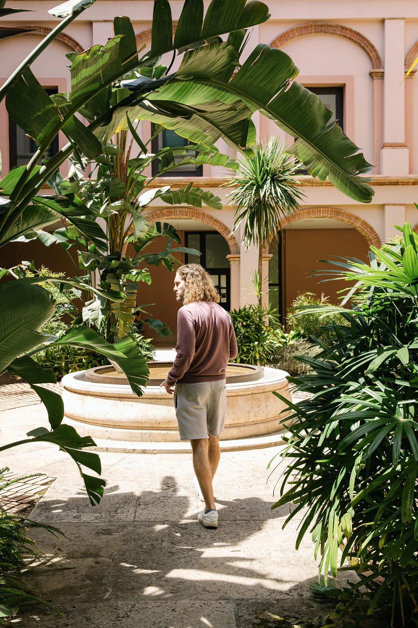A young man with curly hair, wearing a brown sweatshirt, beige shorts, and white sneakers, standing near a stone fountain in a courtyard filled with lush green tropical plants. Behind him is a pink building with arched windows and decorative brickwork.