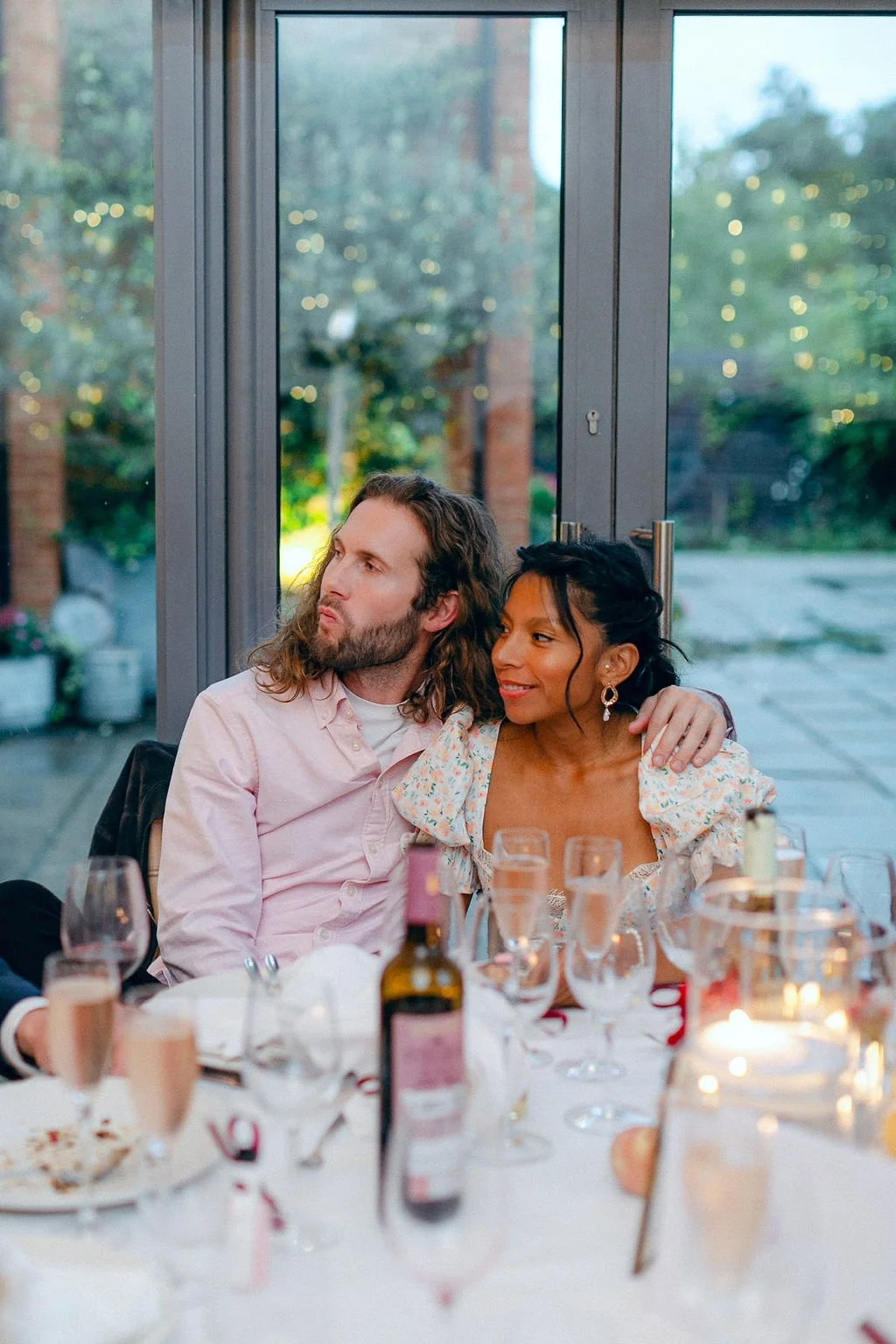 A man with long, curly hair and beard, wearing a light pink shirt, hugging a woman with dark hair and earrings, dressed in a floral dress, sitting at a table with drinks and food during a gathering in a bright room with large windows showing a garden outside.