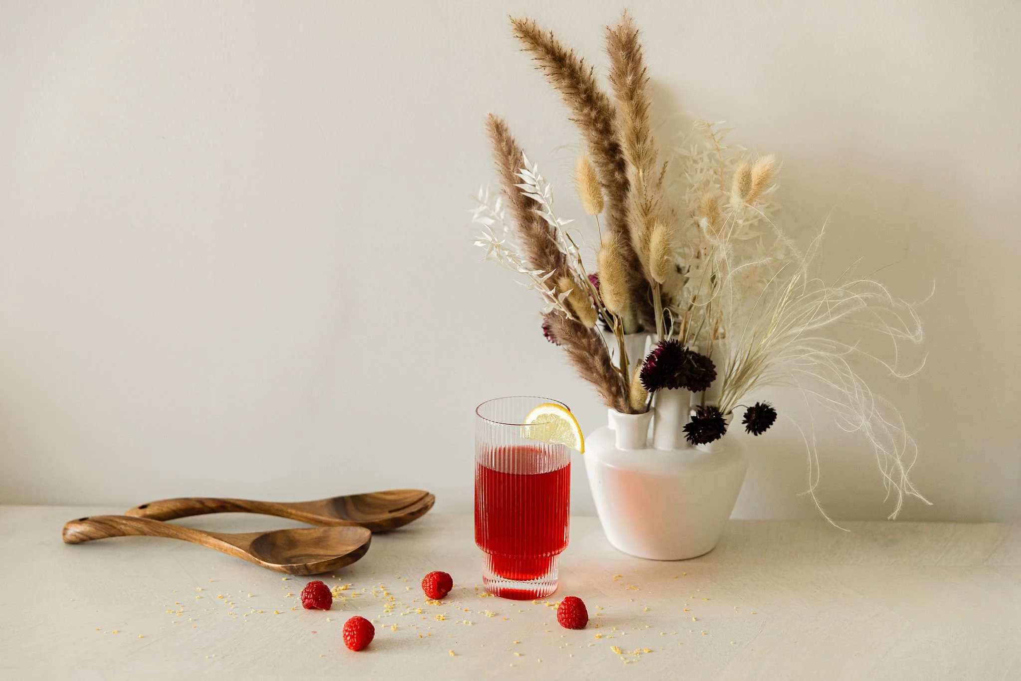 A red glass of juice with lemon wedge, a white vase with dried pampas grass and decorative flowers, and wooden spoons on a light-colored surface with scattered raspberries and yellow sprinkles.
