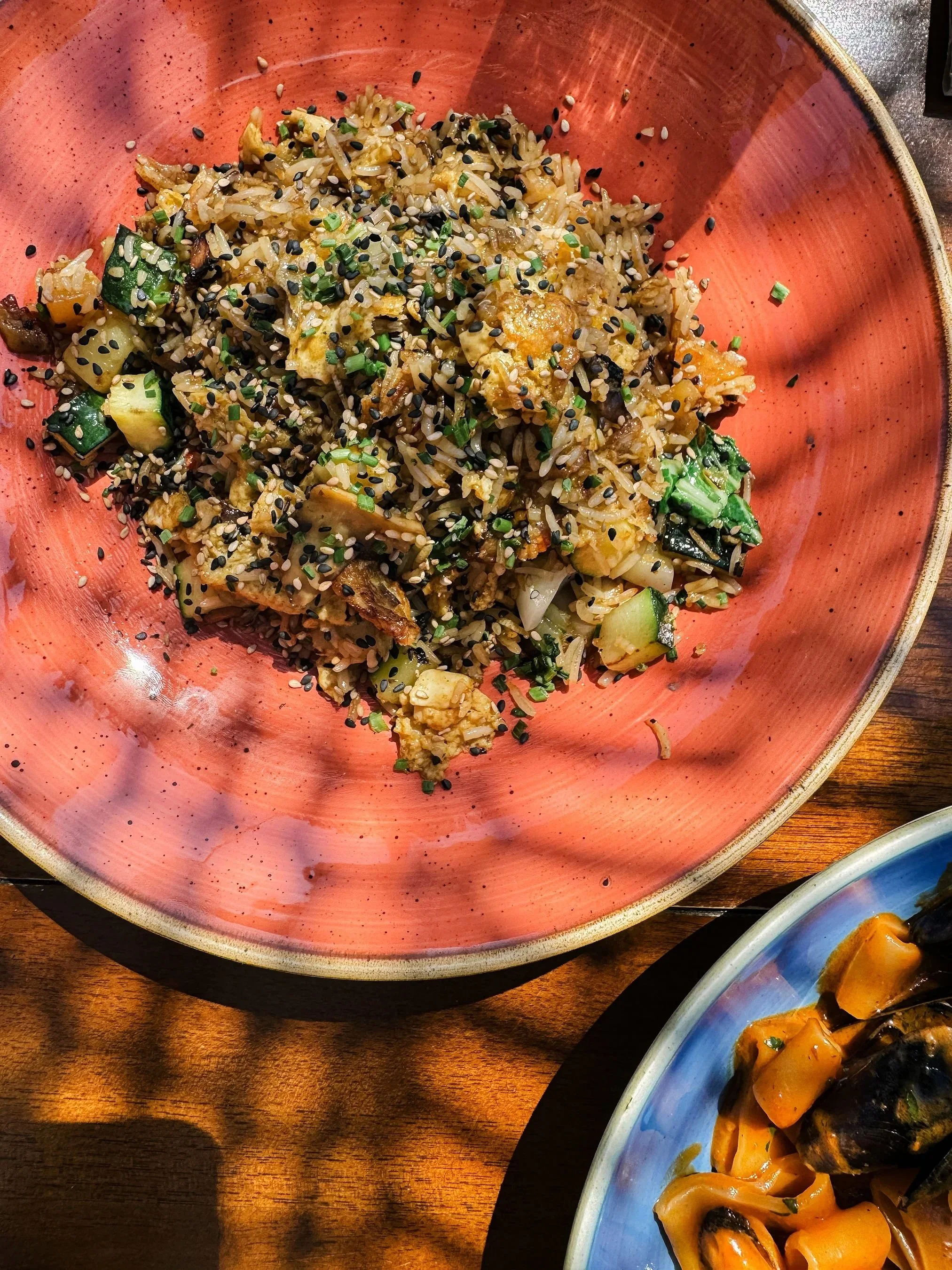 A bowl of fried rice garnished with black and white sesame seeds and chopped green herbs, served in a pink bowl on a wooden table.