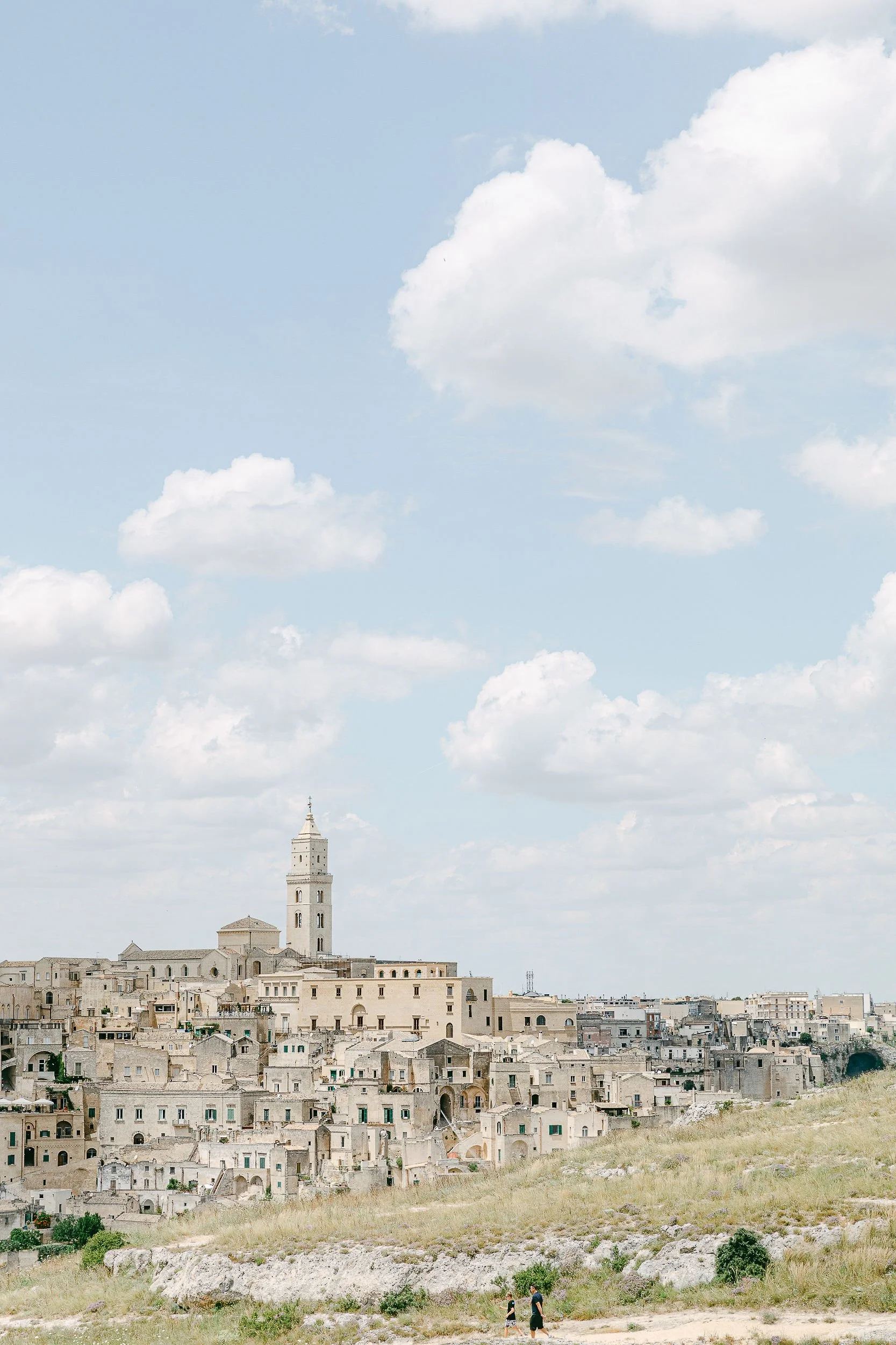 A panoramic view of a historic city with stone buildings and a tall tower under a partly cloudy sky, with two people walking along a grassy path in the foreground.