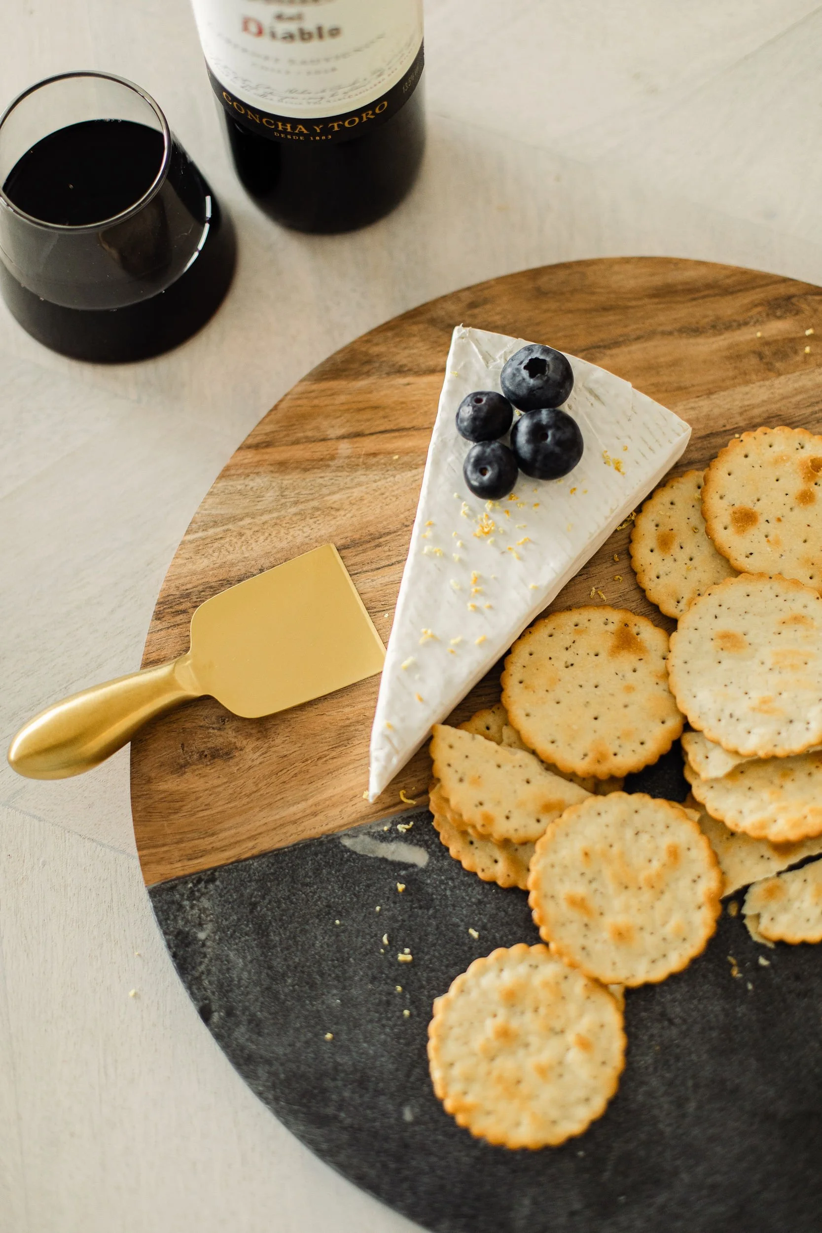 A cheese wheel topped with blueberries, slices of round crackers, and a gold-handled cheese knife on a wooden round board.