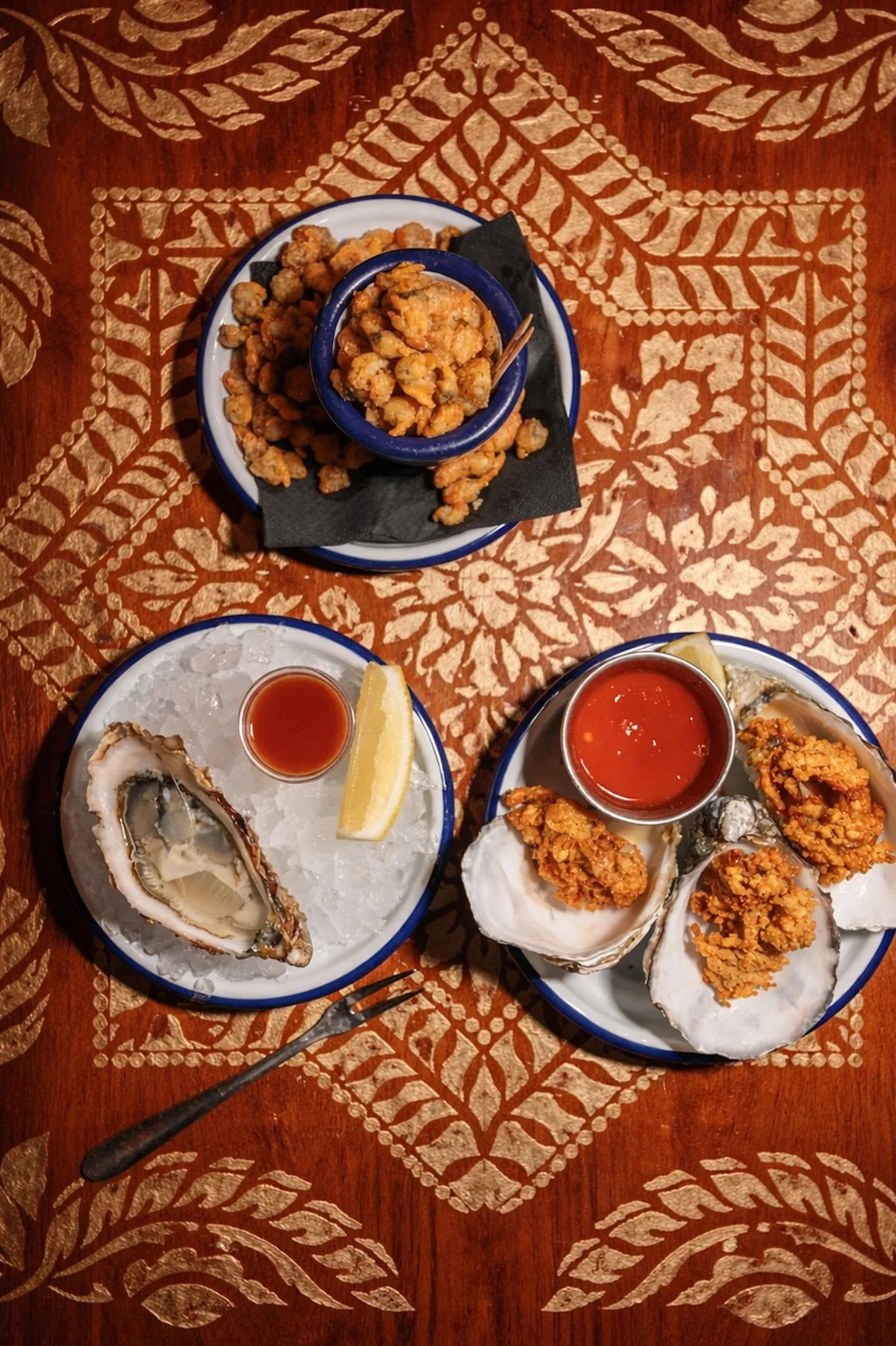 A top-down view of a table with dishes of seafood, including oysters, and fried items, accompanied by sauces and lemon wedges, on a decorative orange tablecloth with gold leaf and floral patterns.