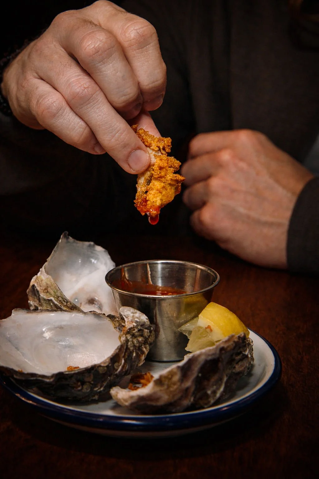 A person holding a piece of fried food over a plate of oysters, a lemon wedge, and a small metal container of sauce.