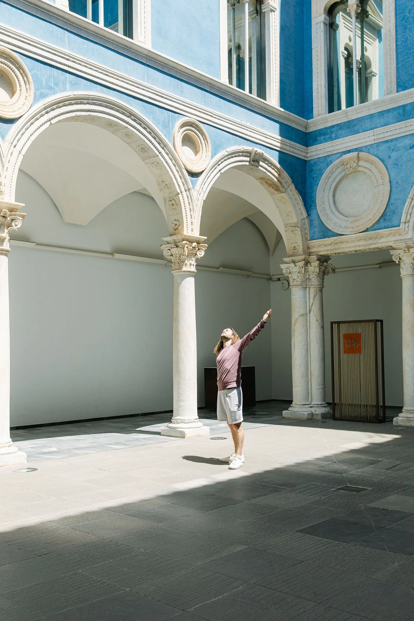 A man with long hair, beard, wearing a brown jacket, white shorts, and white shoes, standing and pointing upward in a courtyard with classical architectural features, including arched columns, detailed trim, and a blue and white building facade.