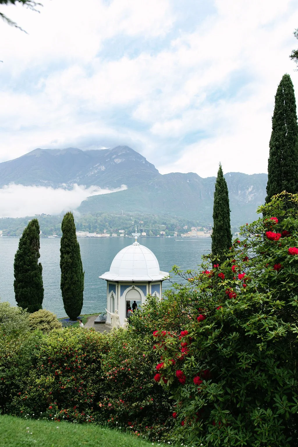 A scenic lakeside view with a picturesque white pavilion, surrounded by tall cypress trees and blooming bushes, with mountains in the background and a cloudy sky overhead.