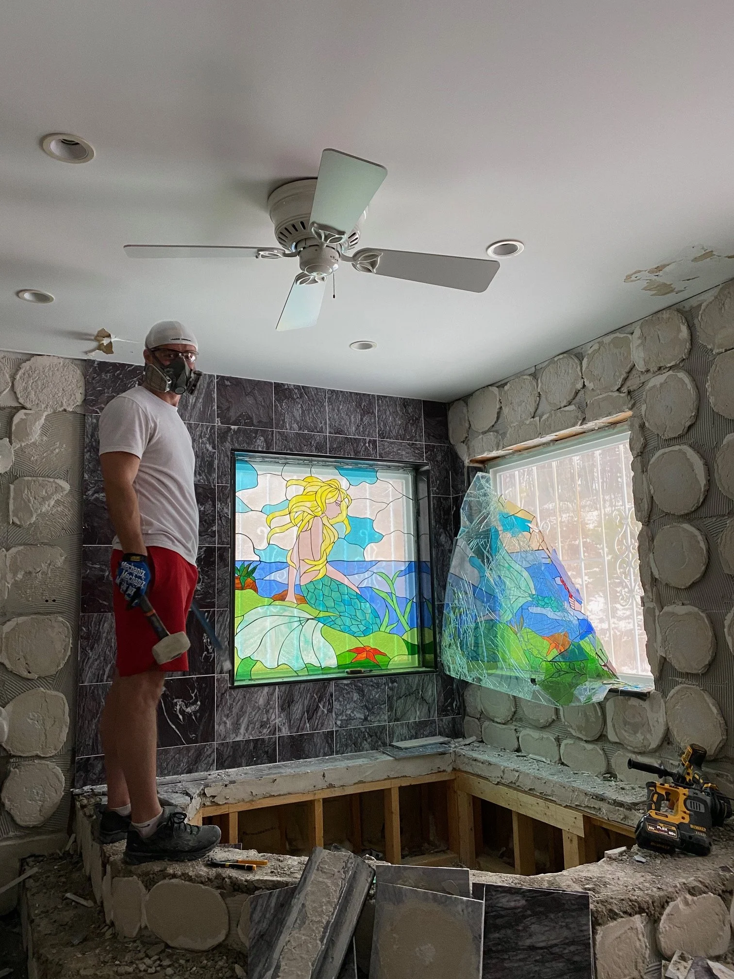 Igor in respirator mask with sledgehammer, mid-demo of the marble bathroom with mermaid window still in place