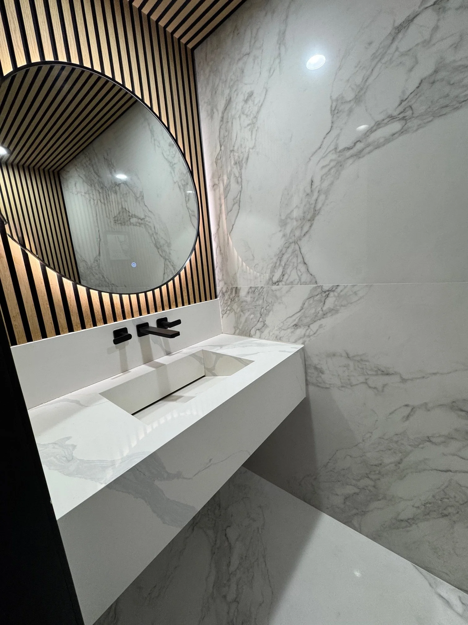 Modern bathroom with marble walls, a white countertop, a black faucet, a wooden slatted accent wall, and a large oval mirror.