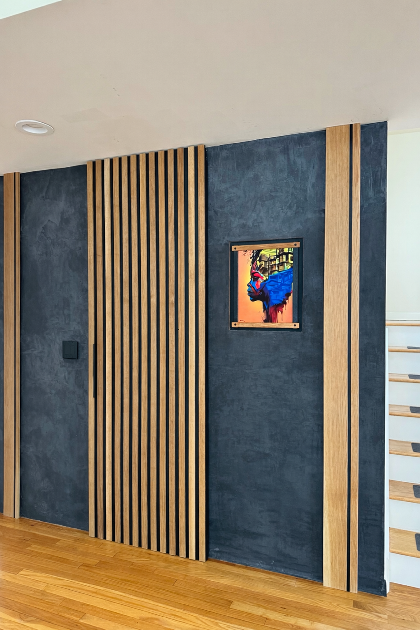 Interior view of a modern room with a dark textured wall, accented by vertical wooden slats, a small colorful framed artwork, and a staircase with light-colored wooden steps and black accents.
