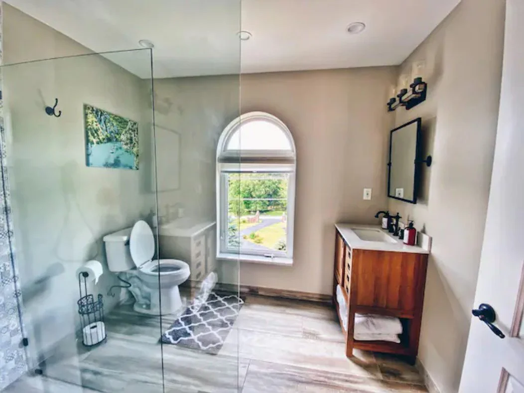 Bathroom with a toilet, a window, a wooden vanity with a sink, a mirror, and a glass shower enclosure.
