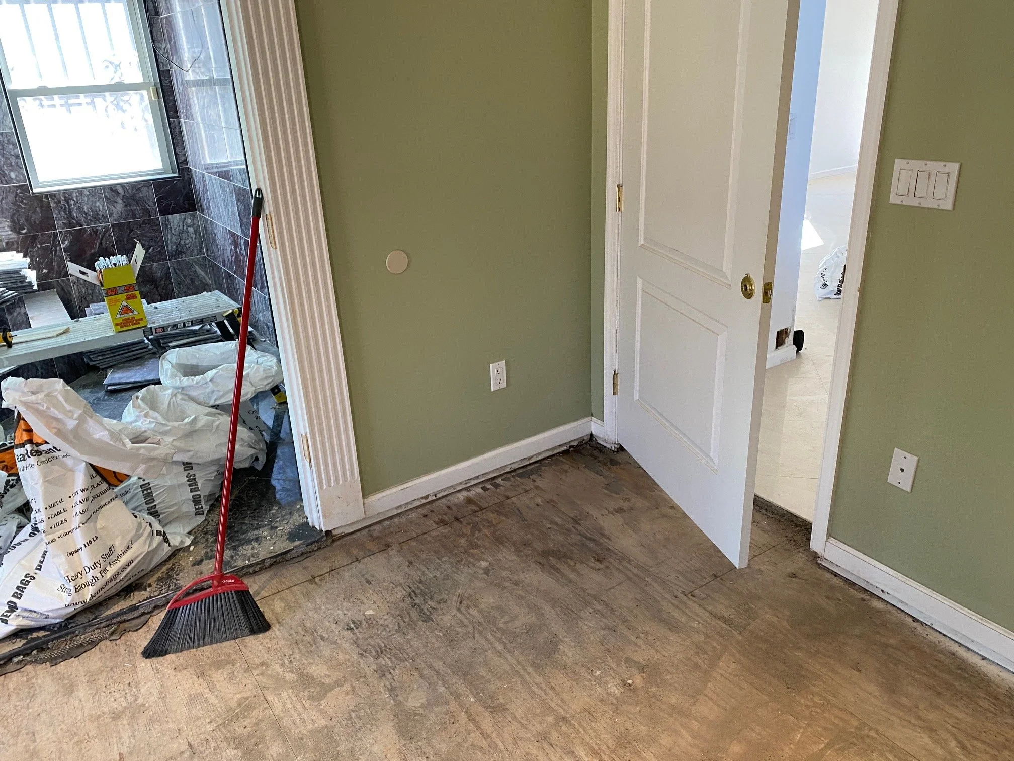 Room undergoing renovation with partially removed hardwood flooring, a broom leaning against the wall, and construction materials near a doorway to a bathroom with black marble tiles.