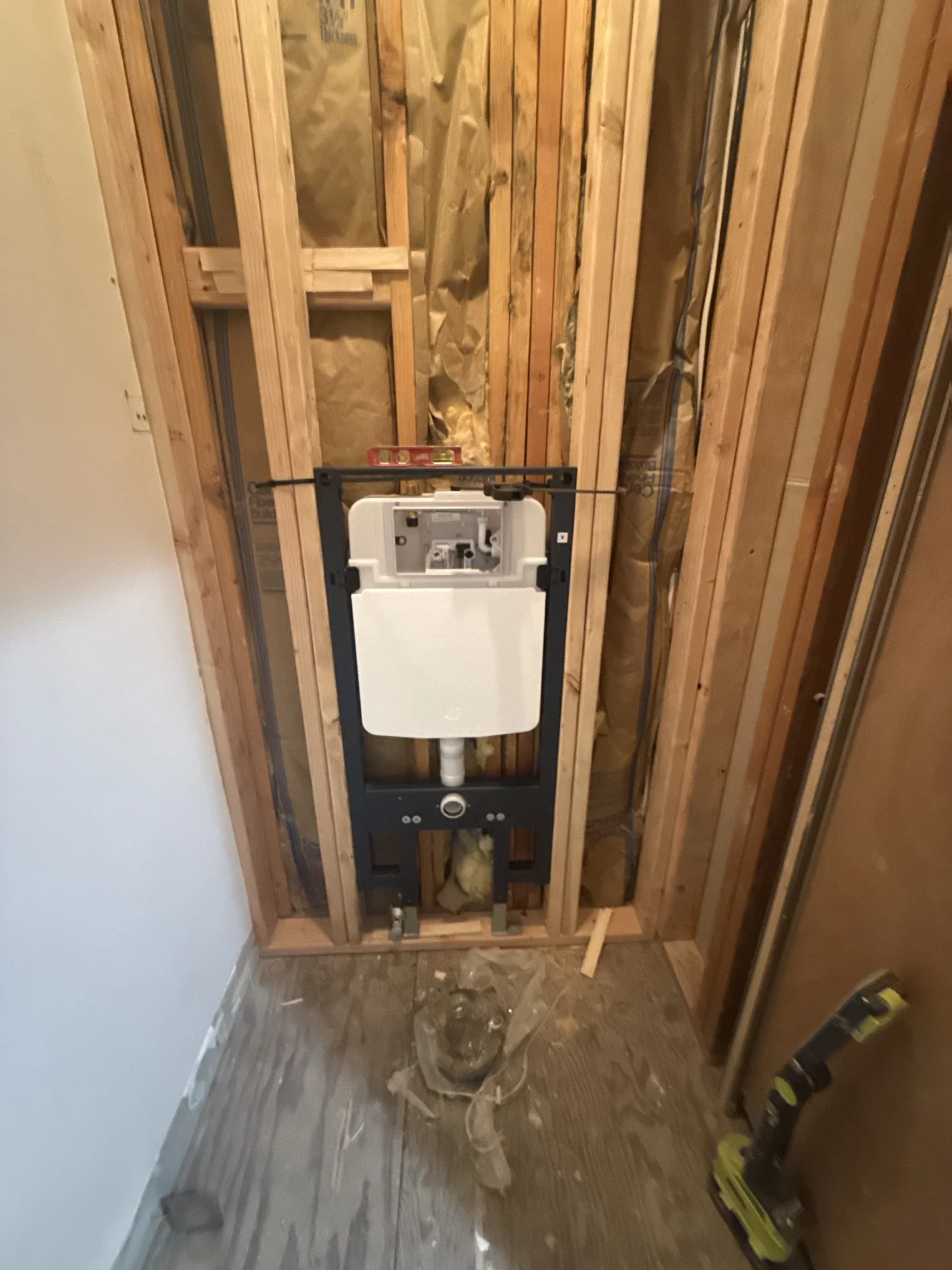 Indoor construction scene with exposed wooden studs and drywall, and a mounted toilet tank framework installed in the wall.