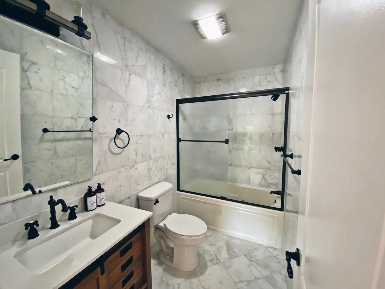 Bathroom with marble walls and floor, black fixtures, a white toilet, vanity with black hardware, mirror, and a tub with a glass shower door.