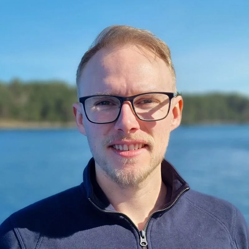 David Remø. En mann med brunt hår, briller, og skjegg smiler mot kamera, med vann og trær i bakgrunnen på en klar, solrik dag.