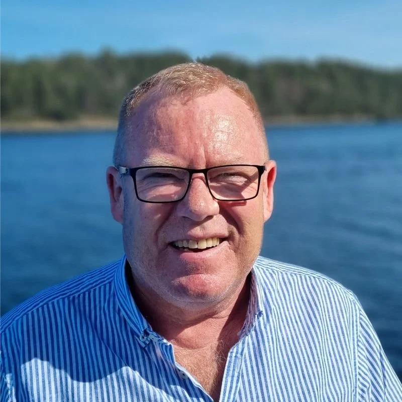 Johnny Remø. En mann med brunt hår og briller, iført en blå stripete skjorte, med grønne trær og sjø i bakgrunnen på en solrik dag.