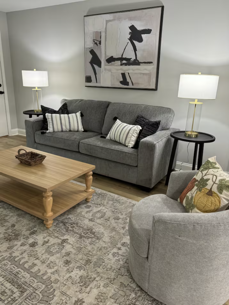 Living room with gray sofa, armchair with decorative pillow, wooden coffee table, two side tables with lamps, abstract wall art, and area rug.