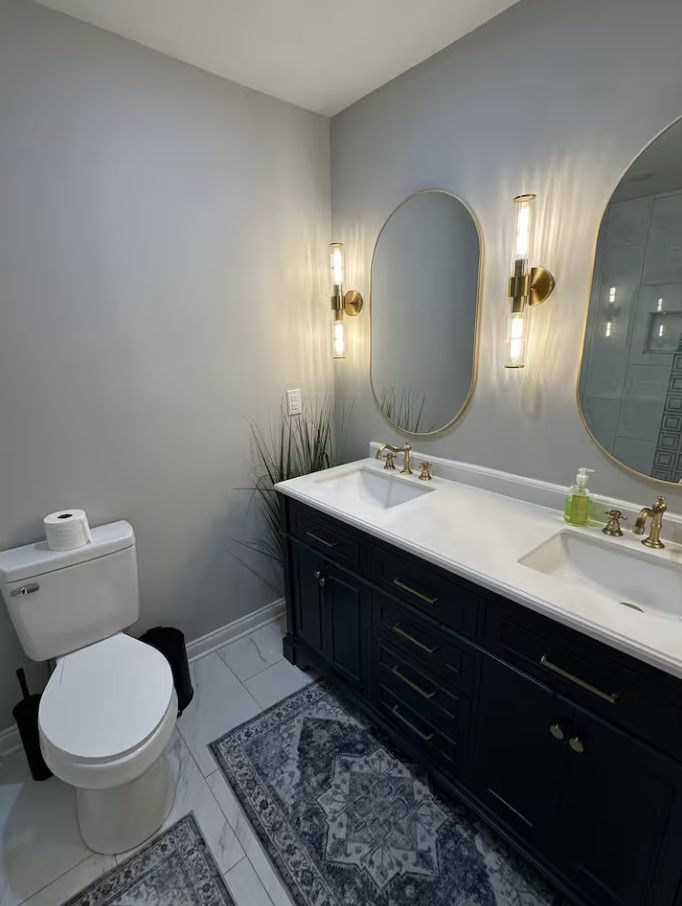 Modern bathroom with gray walls, double sink vanity with dark cabinetry, oval mirrors, gold fixtures, wall-mounted lights, a toilet, and patterned rugs on white tiled floor.