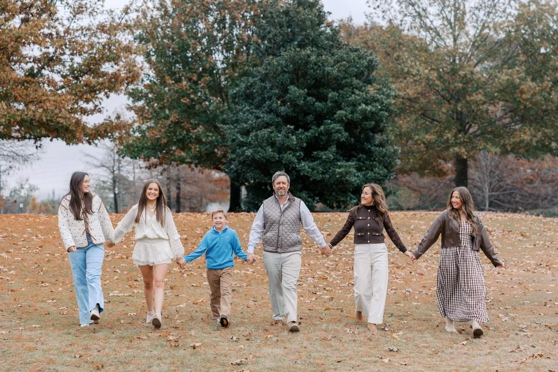 Family walking hand-in-hand through a park during fall, with colorful autumn trees and fallen leaves.