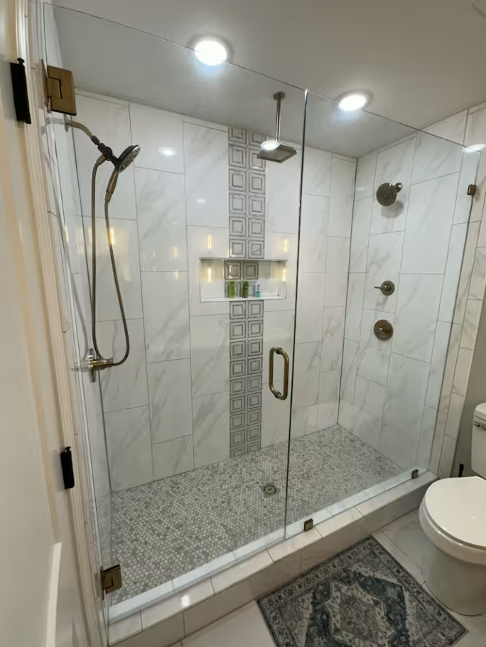 Glass-enclosed walk-in shower with marble tiles, built-in niche with toiletries, overhead rain showerhead, and multiple body spray nozzles, next to a toilet and a decorative rug.