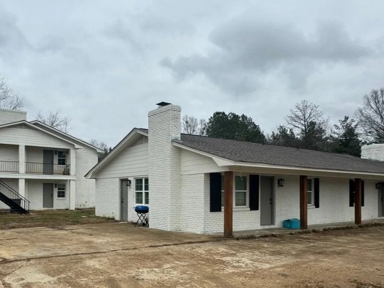 White brick house with a chimney, multiple windows, and a covered porch supported by wooden posts, located in a yard with bare soil.