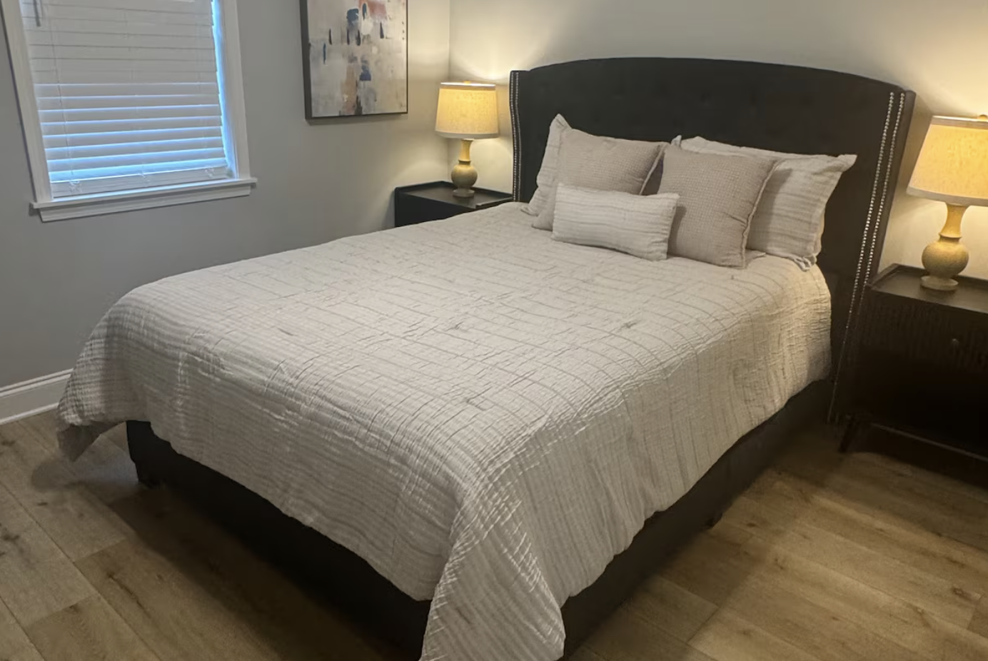 A neatly made bed with beige bedding and multiple pillows in a bedroom with wooden floors, two nightstands with lamps, a window with blinds, and a painting on the wall.