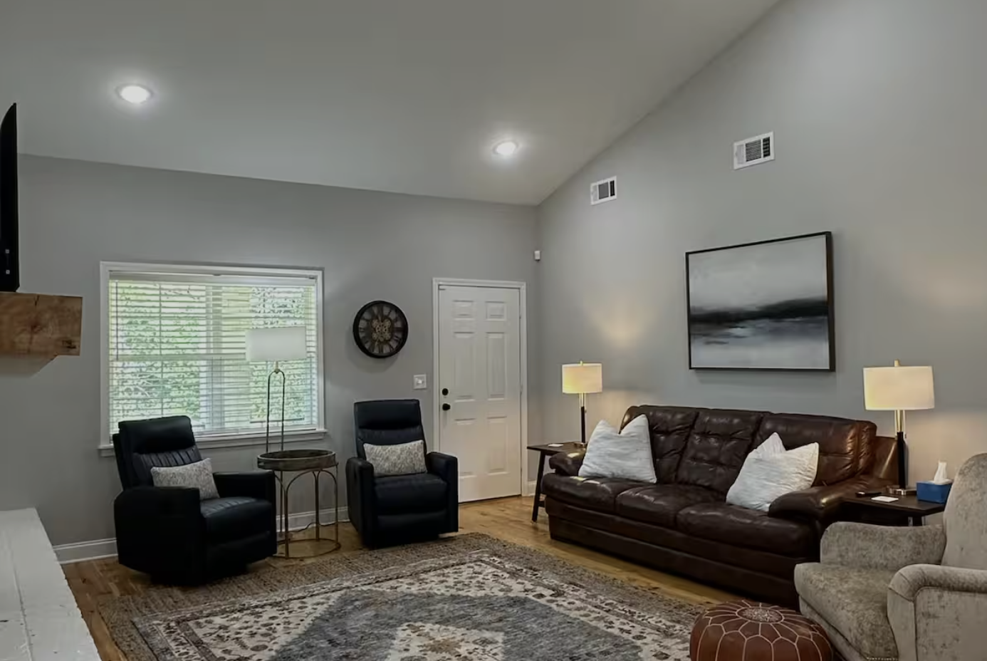 Living room with two black armchairs, a brown leather sofa, an off-white armchair, two side tables with lamps, a patterned area rug, wall art, a window with blinds, and a ceiling with recessed lighting.