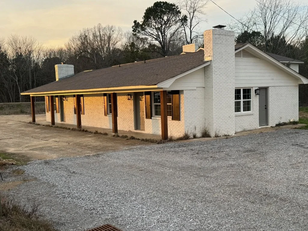 A house with white brick exterior walls, a brown shingled roof, and a chimney. The house has a long covered porch with wooden posts and shutters on the windows. The surrounding area has gravel and dirt, with some trees in the background.