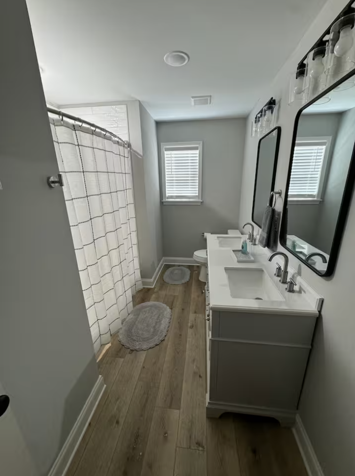A modern bathroom with a white vanity, black-framed mirrors, and a glass jar storage on top. There is a shower with a checkered curtain, a small window with blinds, hardwood flooring, and a gray bath mat.