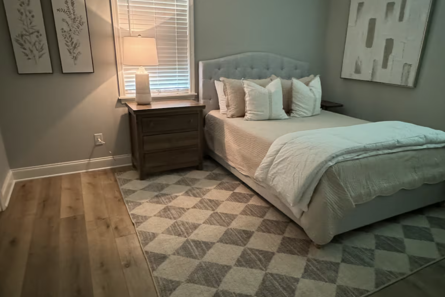 A cozy bedroom with a bed, multiple white pillows, a beige blanket, a wooden nightstand with a white lamp, a window with blinds, and wall art above the bed. The floor has a mix of wooden flooring and a patterned rug.