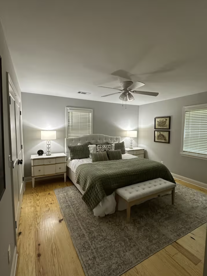 A cozy bedroom with a white tufted headboard, gray bedding, and multiple pillows. Two white nightstands with lamps on either side, a bench at the foot of the bed, and framed artwork on the wall. Hardwood floors with a gray area rug, two windows with 