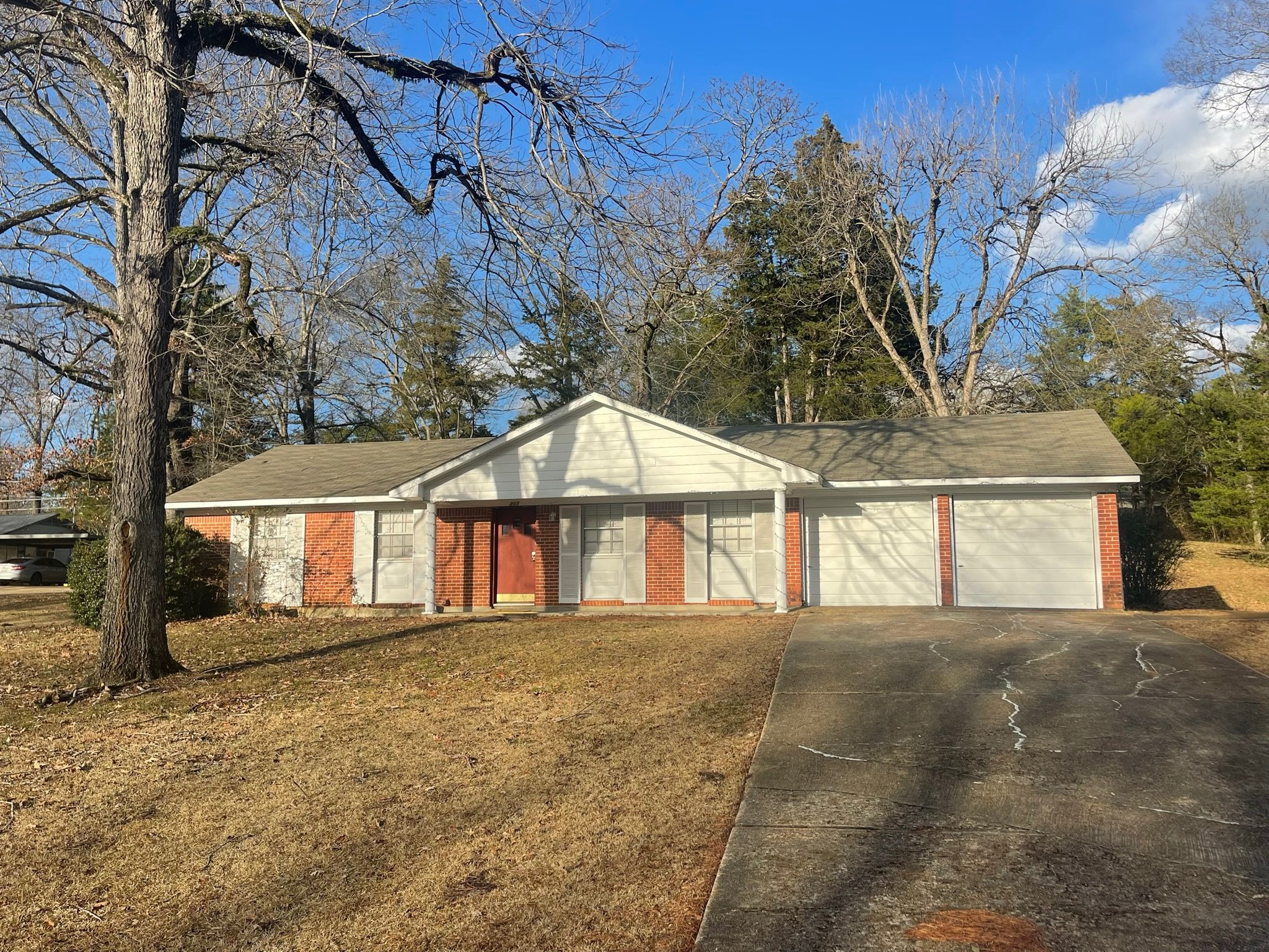 A house with white siding and a gray shingled roof under a bright blue sky. There are tall, leafless trees behind the house, with one large tree to the left of the house. The front yard has patchy grass and fallen leaves, with a concrete driveway on the right side.