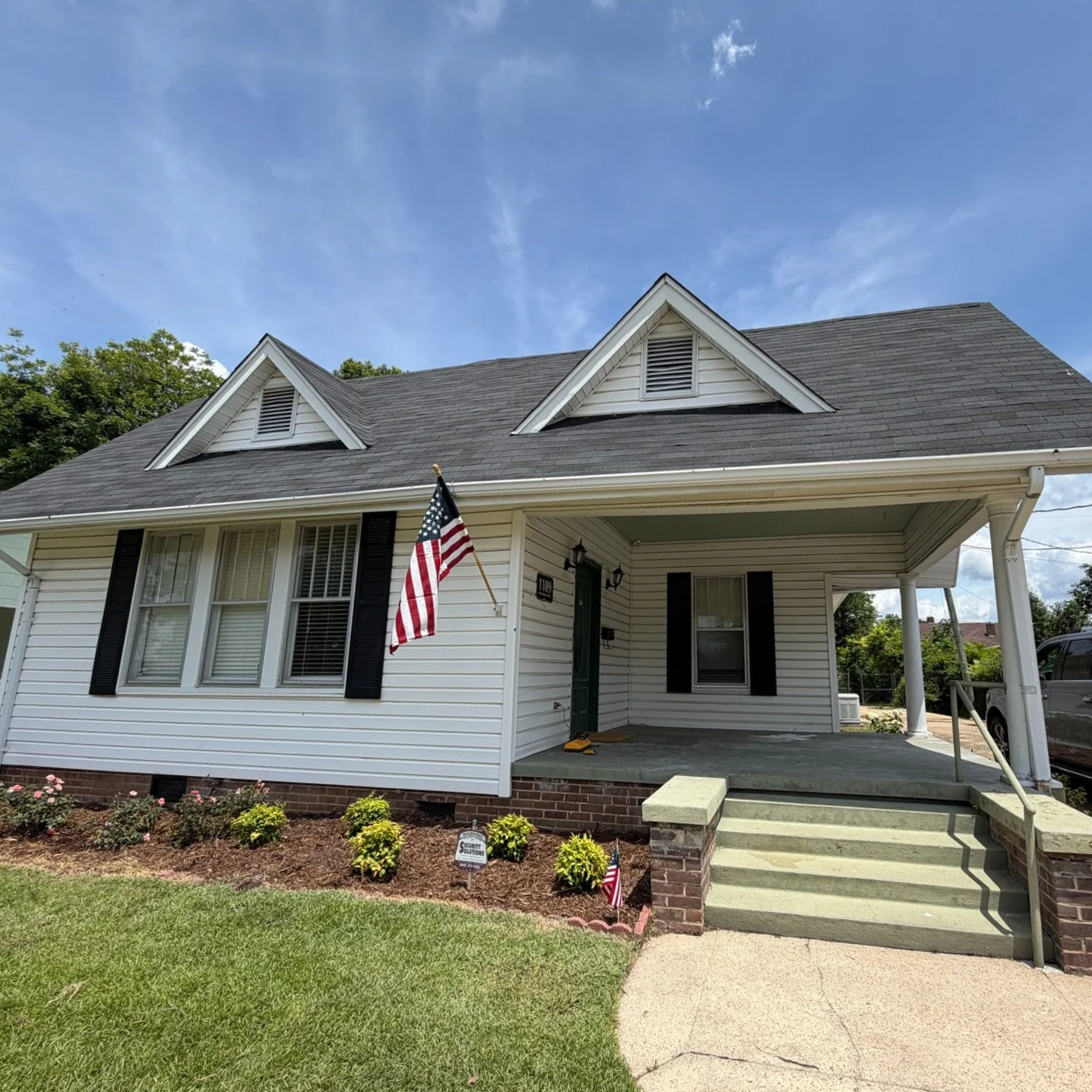 A white house with black shutters and a green front door, American flag hanging near the entrance, front porch with steps, small garden with shrubs and flowers, and a blue sky with some clouds.
