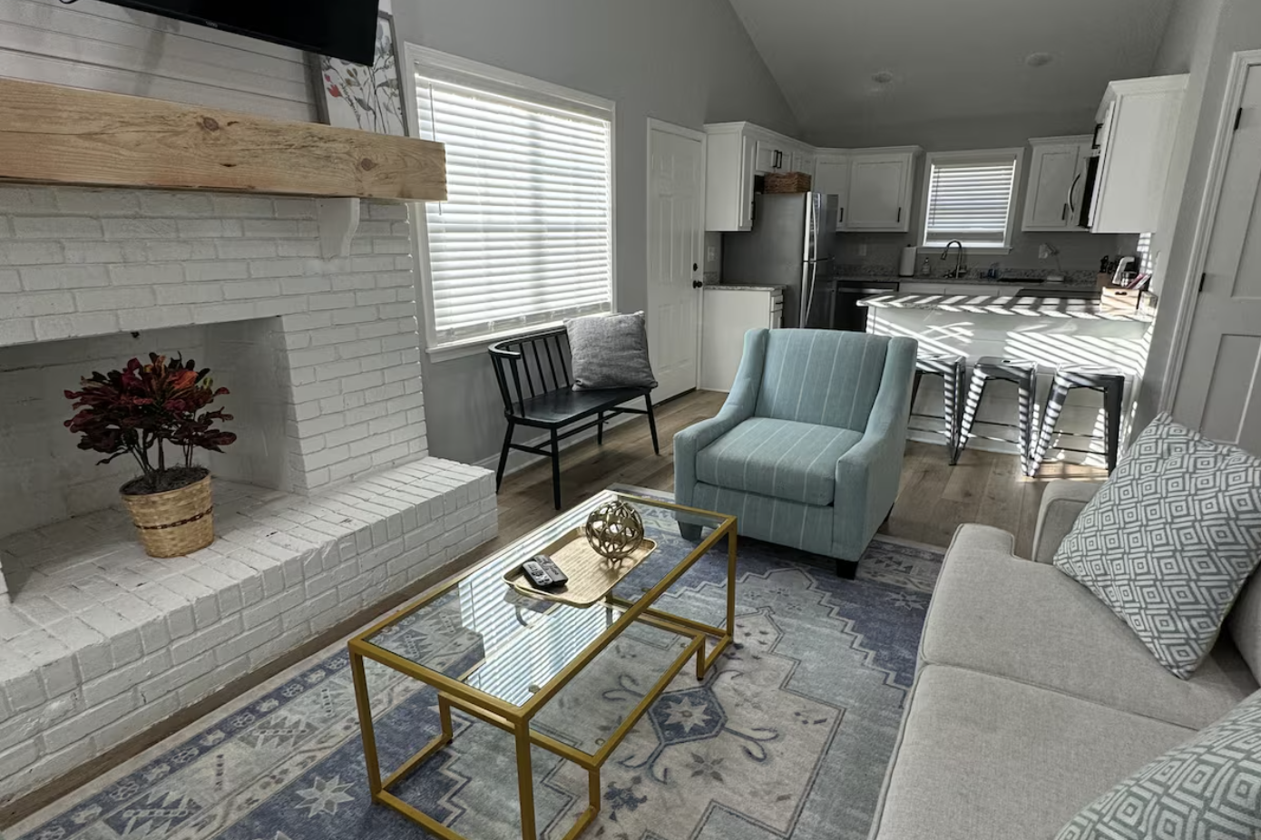 A cozy living room with a white brick fireplace, a flat-screen TV above it, a glass-top coffee table with a decorative sphere, a light blue armchair, a beige sofa with patterned cushions, and a kitchen in the background with white cabinets and a brea