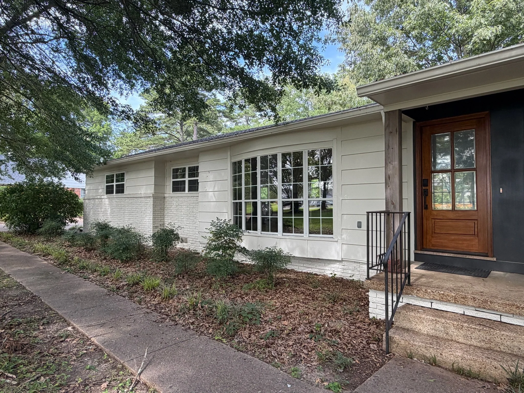 Front view of a house with white siding, large windows, and a wooden door, surrounded by trees and bushes, with a sidewalk in front.