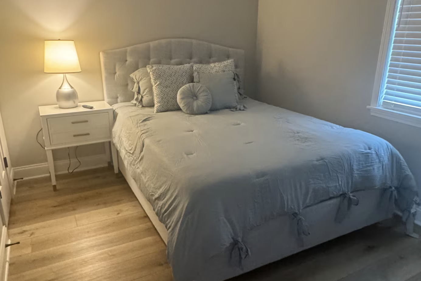 A neatly made bed with a white tufted headboard, several pillows including patterned and round ones, a white side table with a lamp, living room window with blinds, and hardwood flooring.