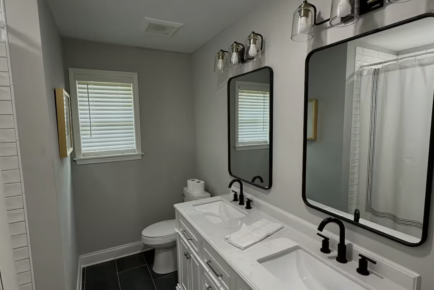 A modern bathroom with a double sink vanity, black faucets, two rectangular mirrors, black light fixtures, a window with white blinds, a toilet, and a shower with a white curtain.