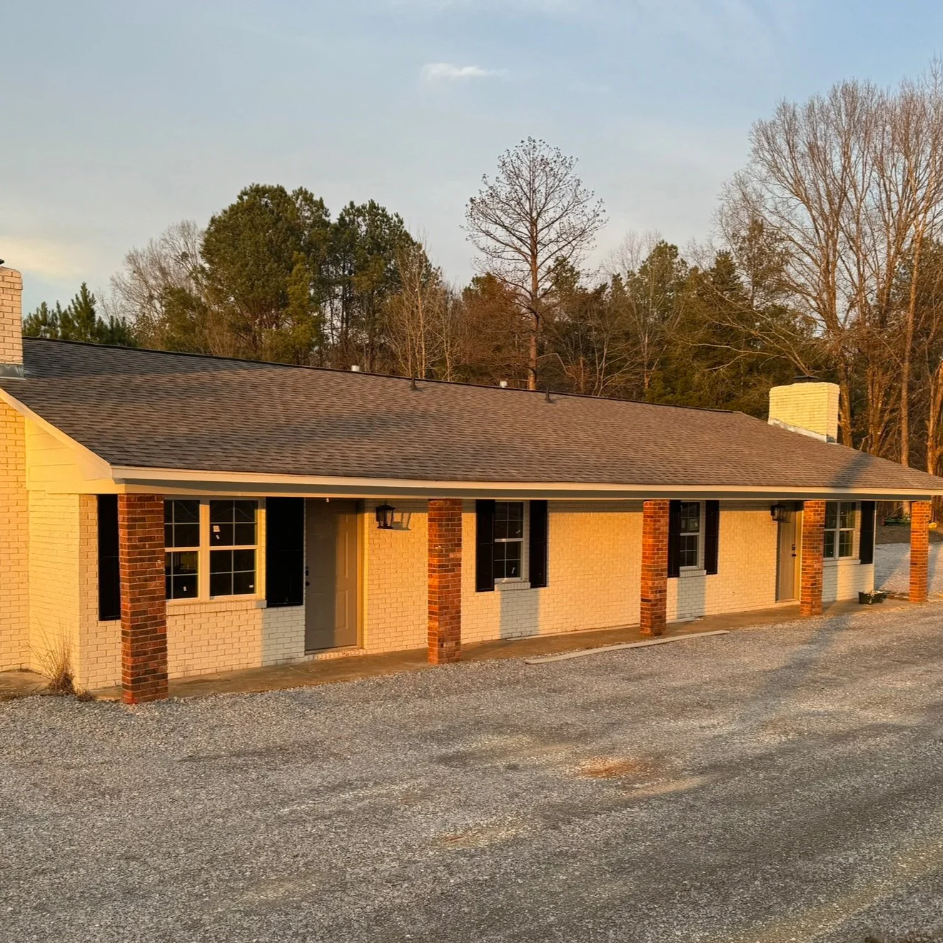 A single-story brick building with black shutters, a gray shingled roof, and an outside covered walkway with supporting brick columns. The building is illuminated by sunlight, with a gravel parking lot in the foreground and trees in the background.