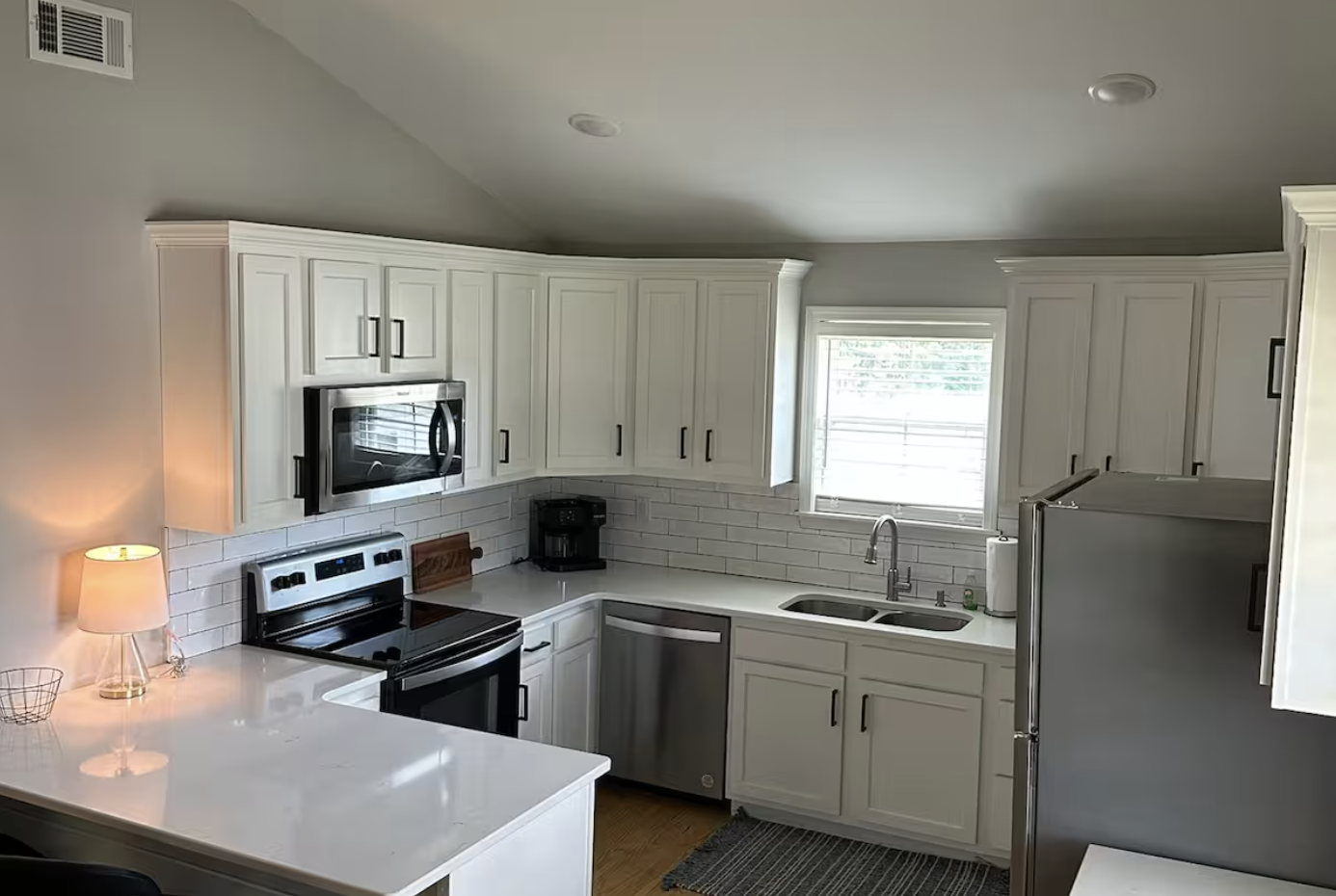 Modern kitchen with white cabinets, stainless steel appliances, a coffee maker, a window above the sink, and a small table with a lamp.